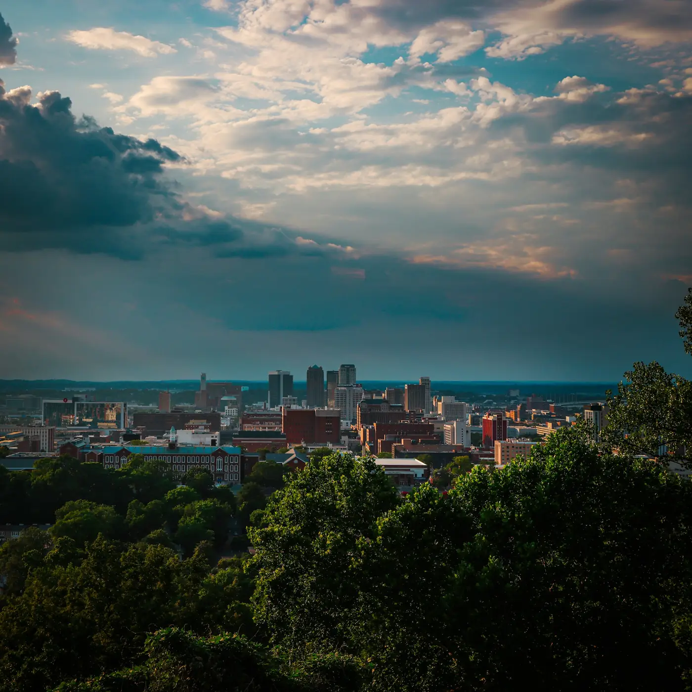 Downtown Birmingham, Alabama skyline beyond a band of green trees under a dramatic sky